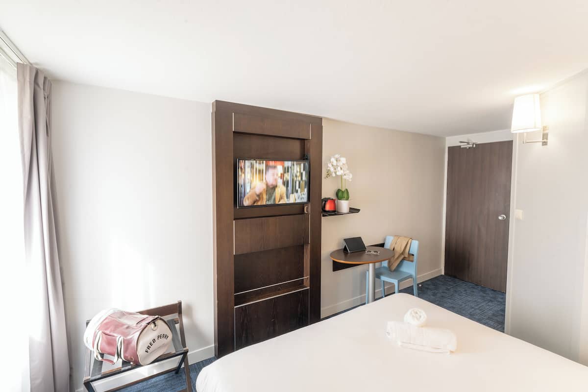 A hotel room with a bed, a desk with a chair, a TV mounted on a wooden panel, a small shelf with a plant, and a door.