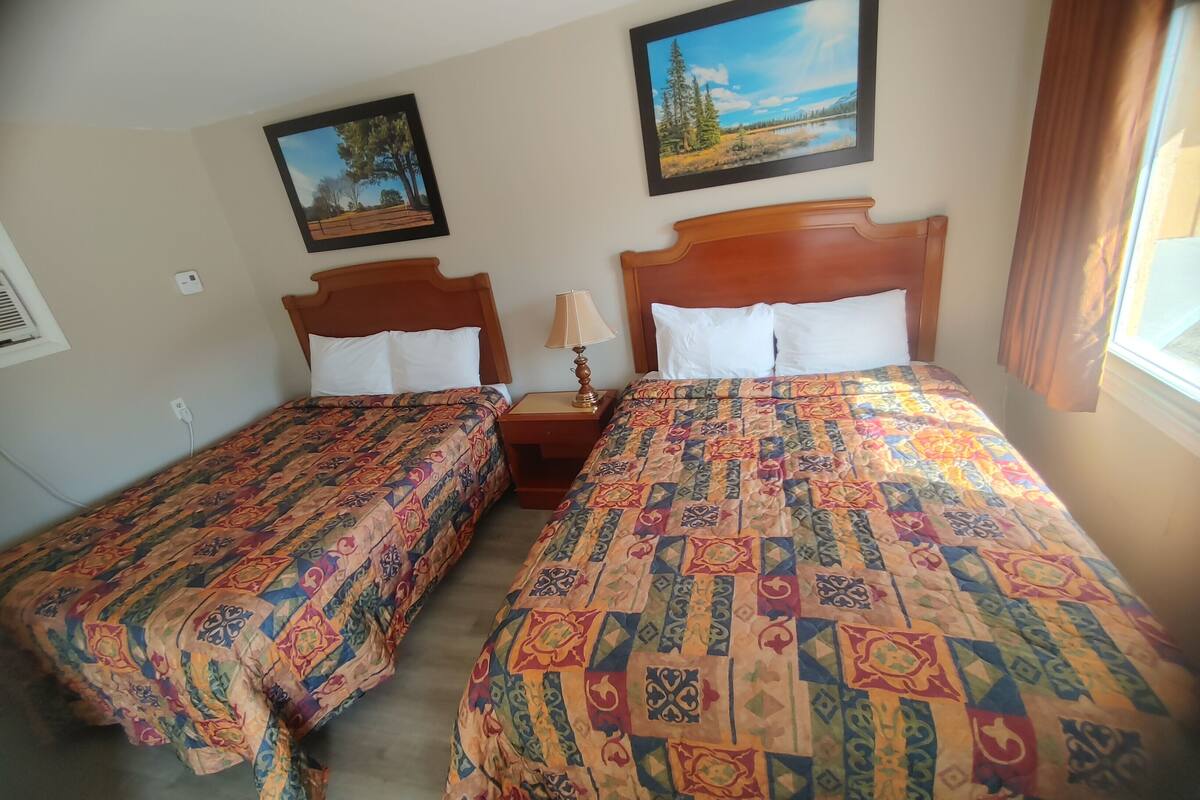 Une chambre avec deux lits, chacun recouvert d’une couvre-lit à motifs et agrémenté d’oreillers blancs. Un tableau est accroché au mur au-dessus des lits, et une fenêtre avec des rideaux se trouve à droite.