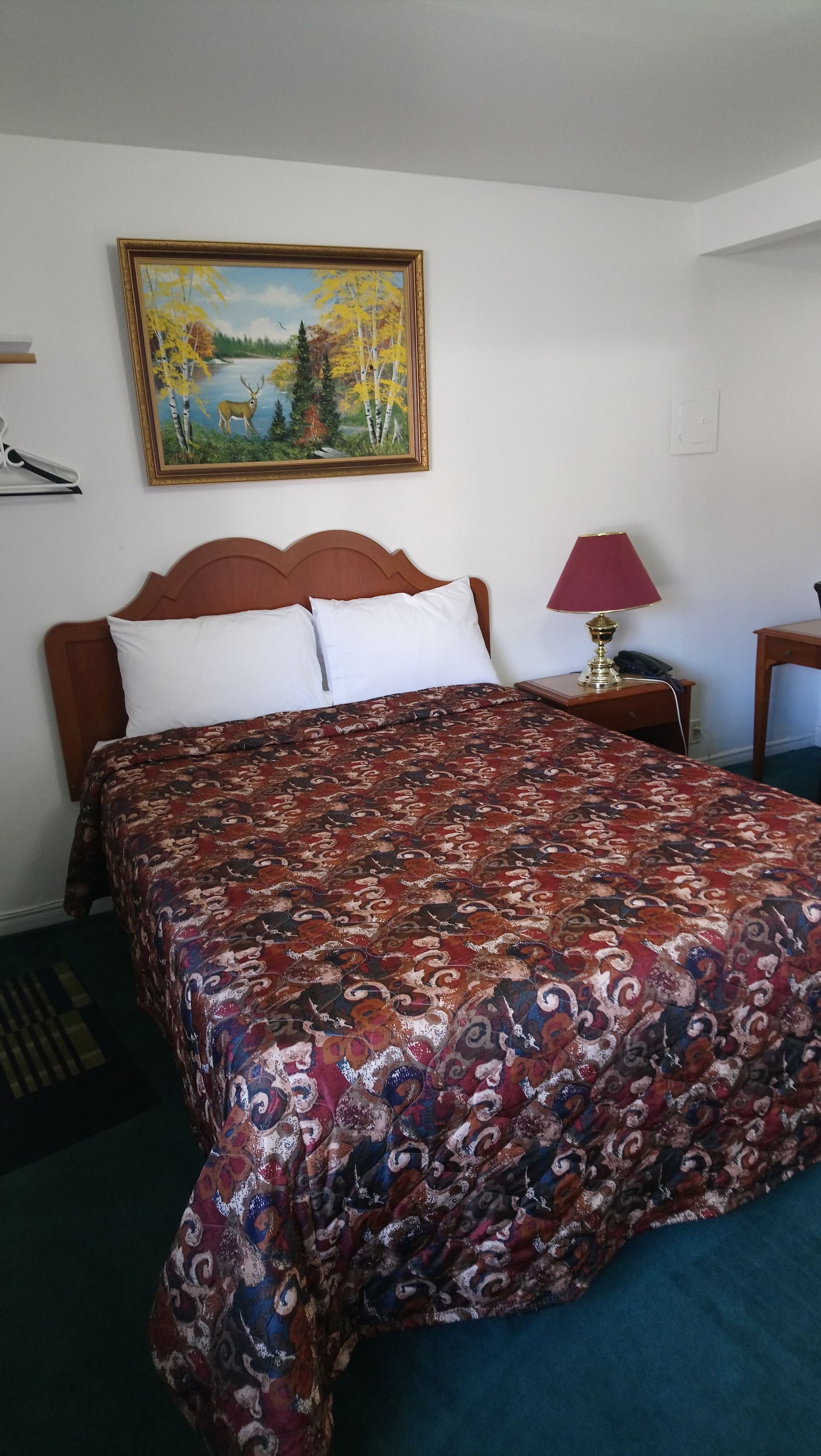 A neatly made bed with a patterned bedspread, a wooden headboard, a painting of a landscape, a bedside lamp, and a telephone on a nightstand.