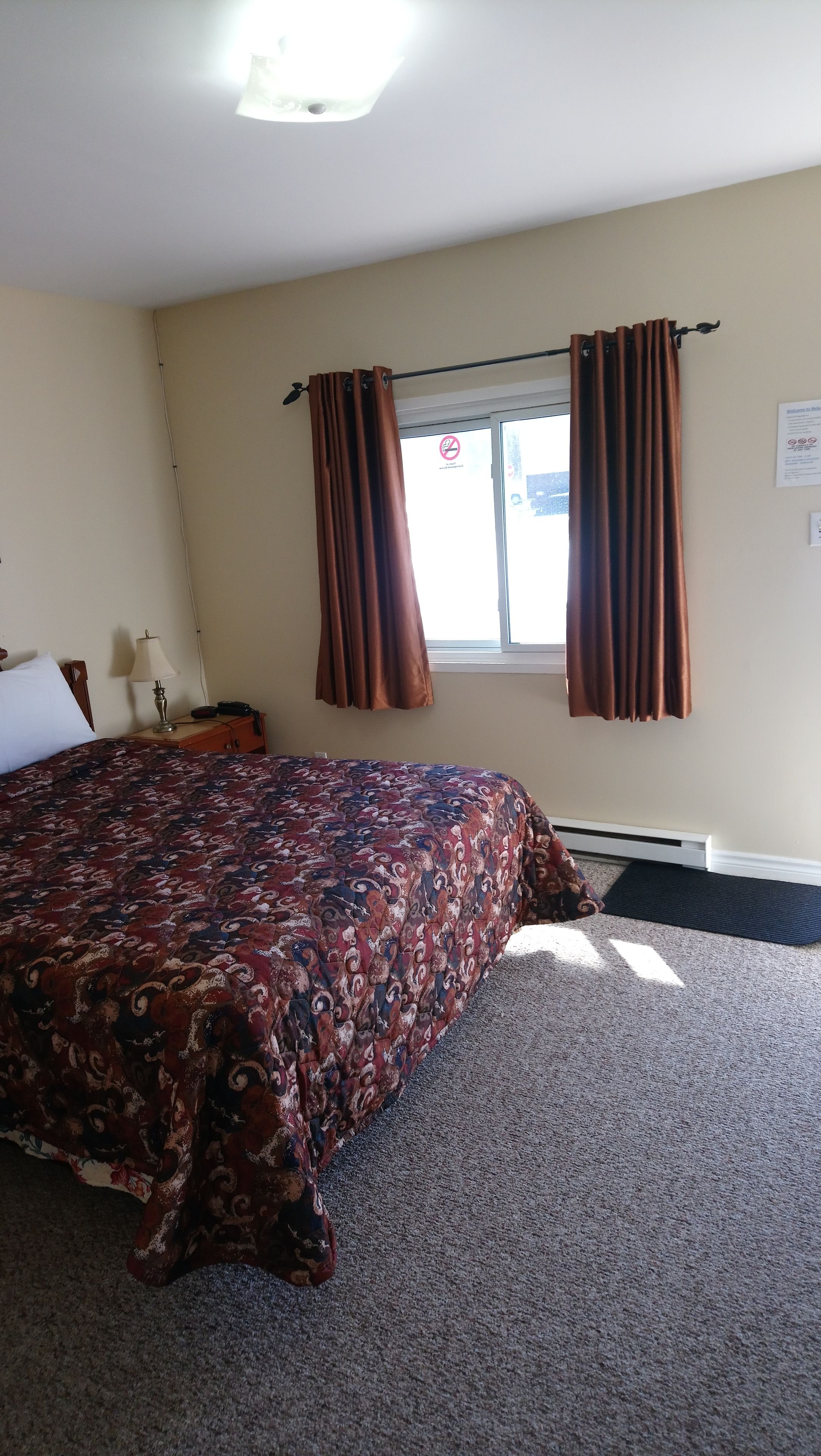 Une chambre d’hôtel équipée d’un lit, d’une table de chevet, d’une lampe, de rideaux, d’une fenêtre et d’un thermostat.