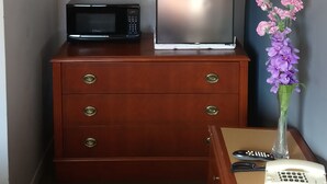 A compact hotel room with a wooden dresser, a refrigerator, a microwave, a vase with flowers, and a bedside table with a phone and remote controls. - Relax Inn Collingwood (Collingwood)
