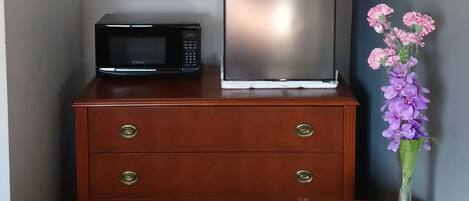 Habitación de hotel compacta con tocador de madera, refrigerador, microondas, un florero con flores y mesita de noche con teléfono y controles remotos.