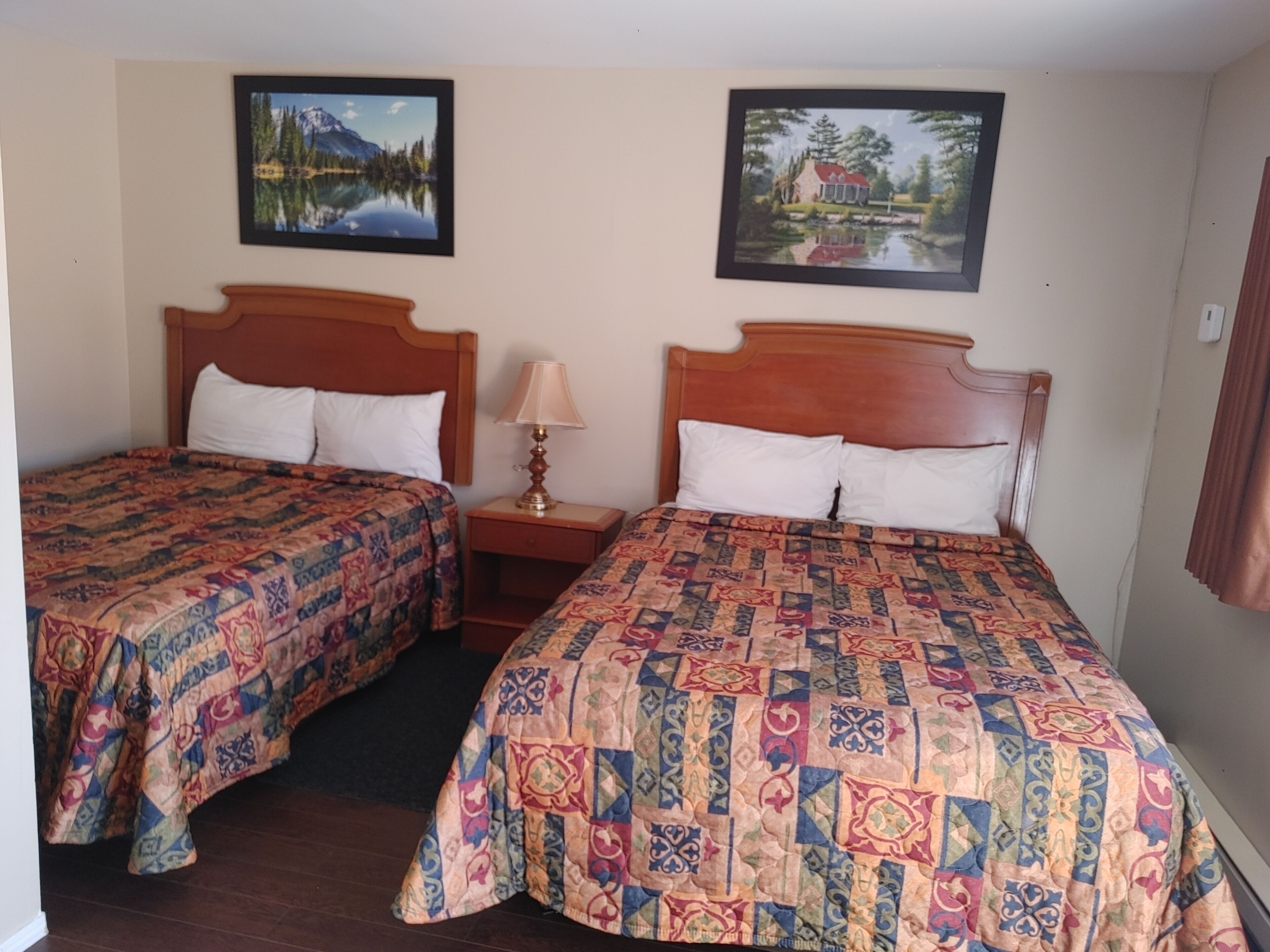 Ein Zimmer mit zwei Betten, jedes mit einem Holzkopfstück und einer gemusterten Bettdecke. An der Wand hängen zwei gerahmte Bilder, eines zeigt eine Berglandschaft und das andere ein Haus am See.
