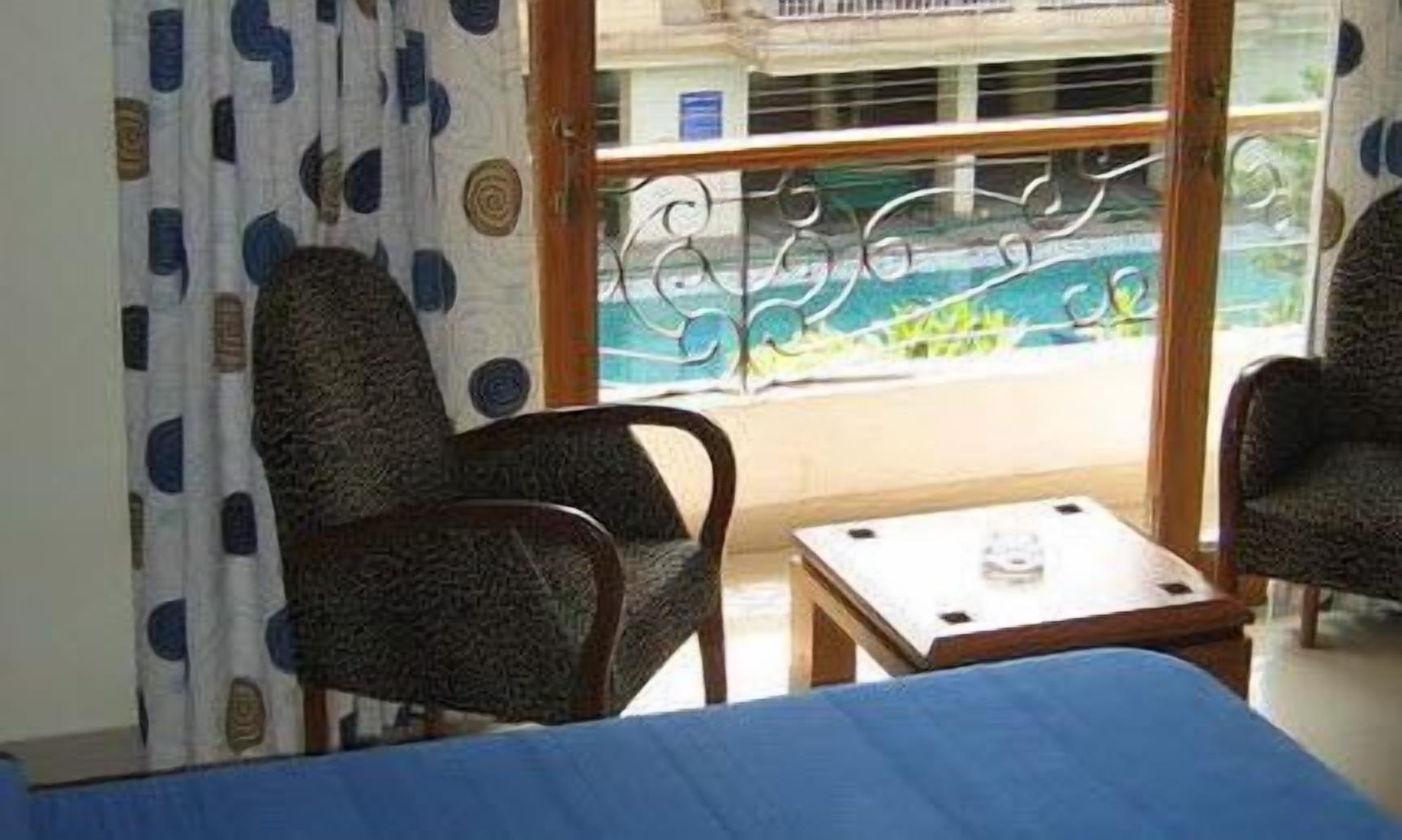 Ein Zimmer mit einem Bett, zwei Stühlen, einem kleinen Tisch und Blick auf einen Pool durch ein Fenster mit Metallgitter.