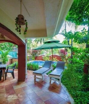 A covered outdoor seating area with two lounge chairs and a table, surrounded by lush greenery and a pool in the background. - Renzo's Inn (Calangute)