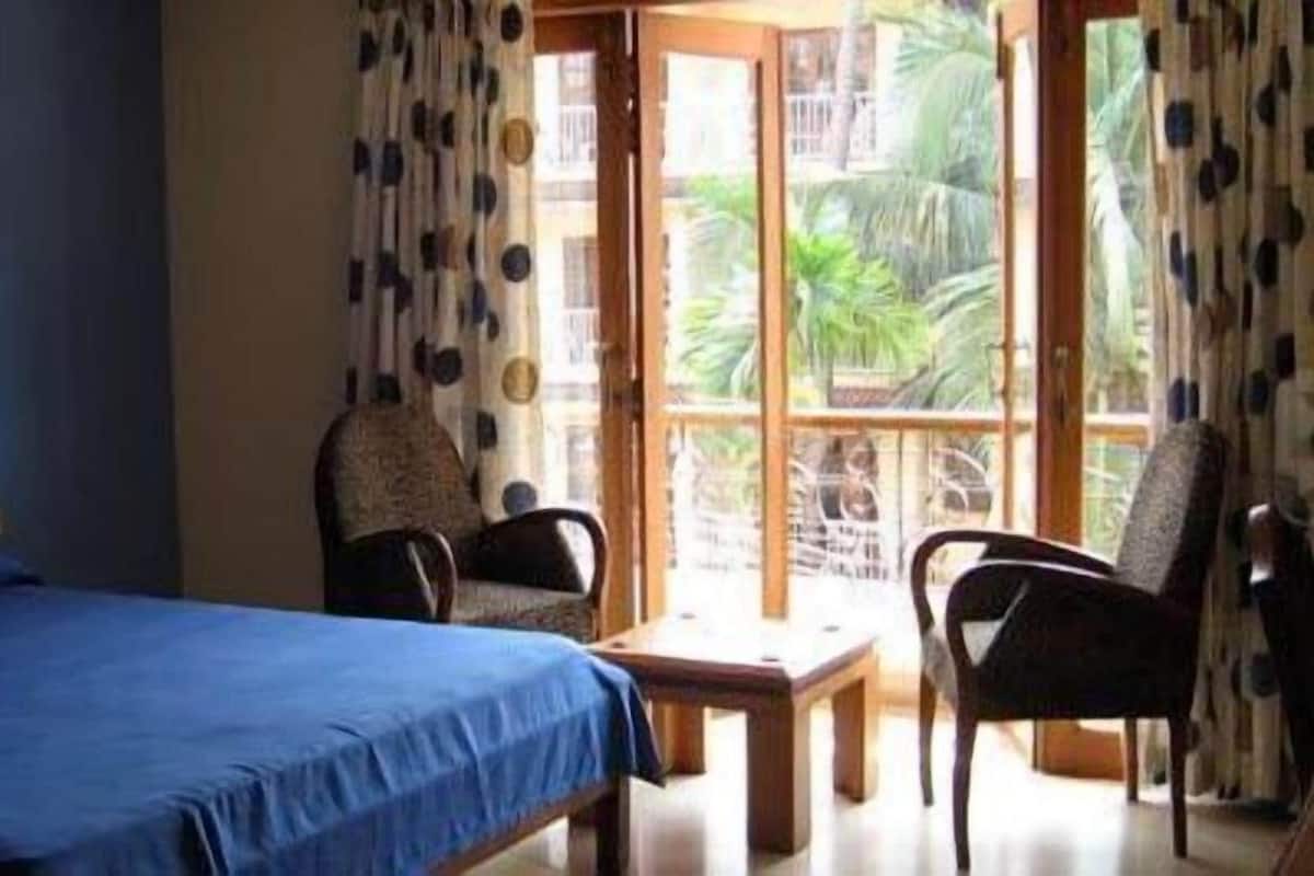 A room with a bed, two chairs, a table, and a large window with a view of greenery.