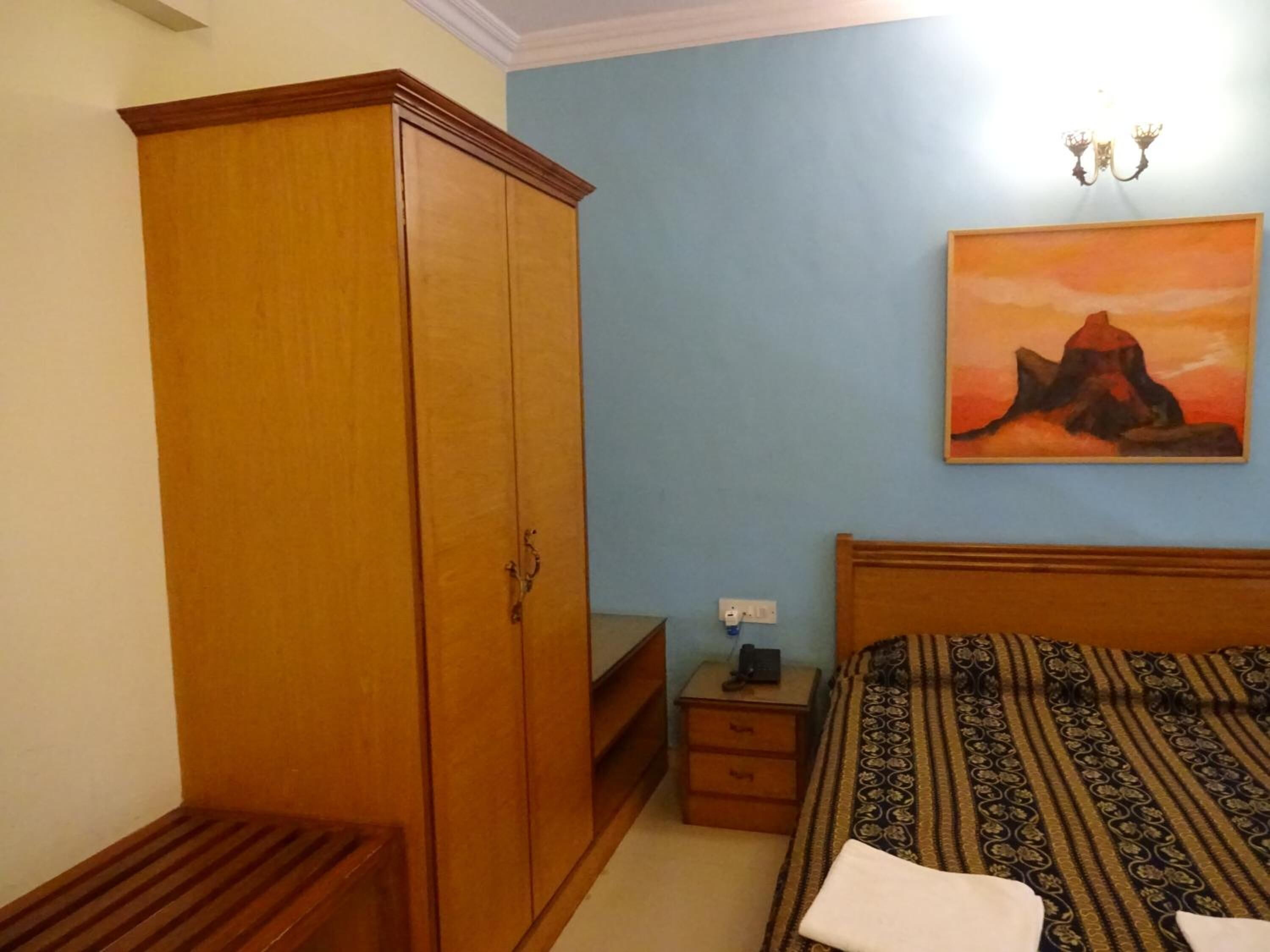 A bedroom with a wooden wardrobe, a bedside table with a lamp, a bed with patterned bedding, and a painting of a mountain on the wall.