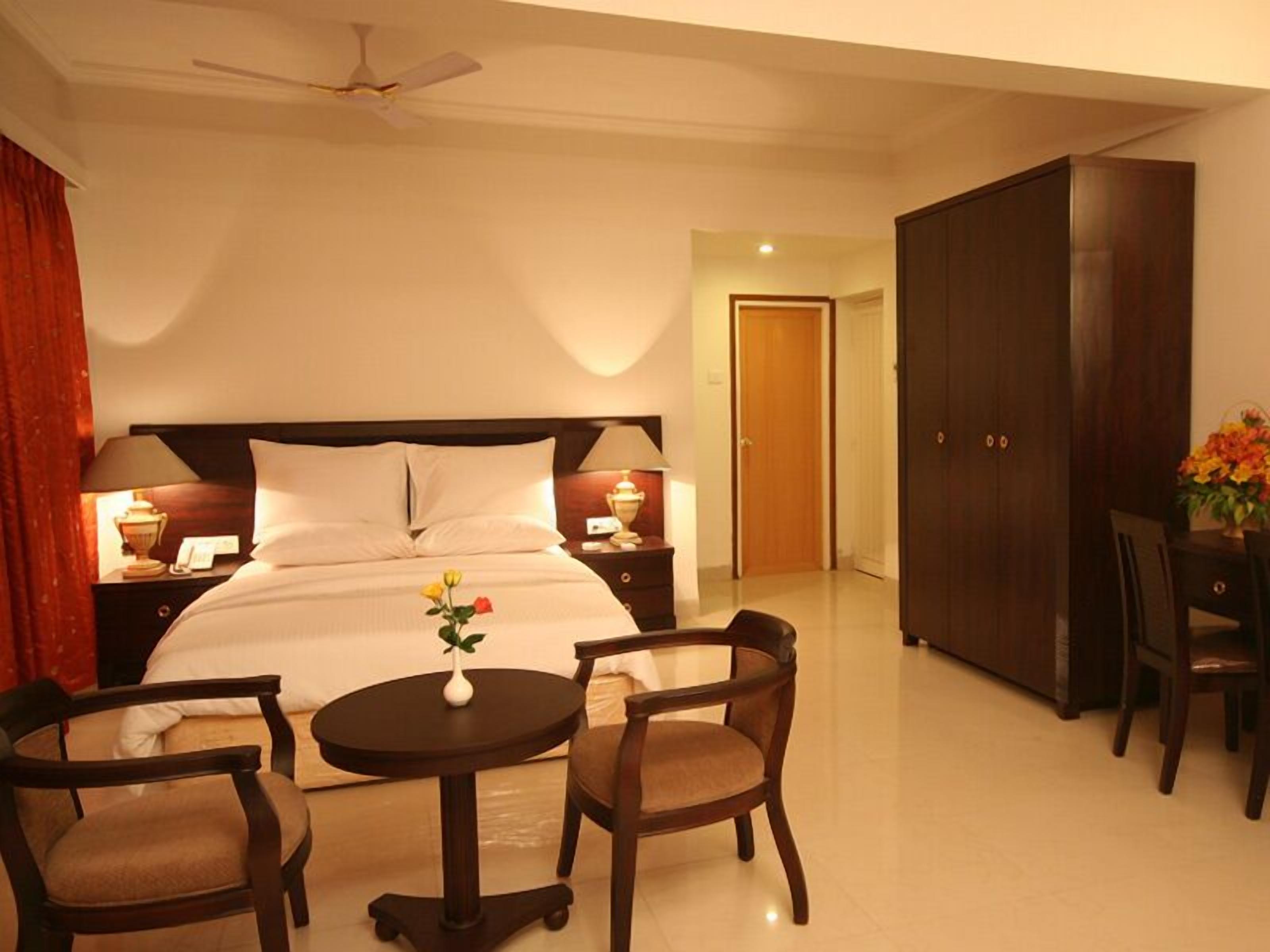 A hotel room with a bed, bedside tables, a desk, chairs, a small table with a vase, and a wardrobe.
