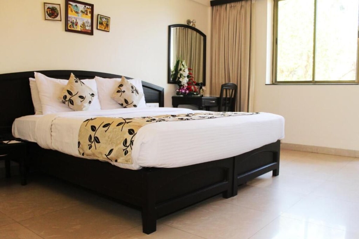 Una cama bien hecha con estructura negra y ropa de cama blanca, una mesita de noche pequeña, una silla, un escritorio con espejo y una ventana con cortinas.