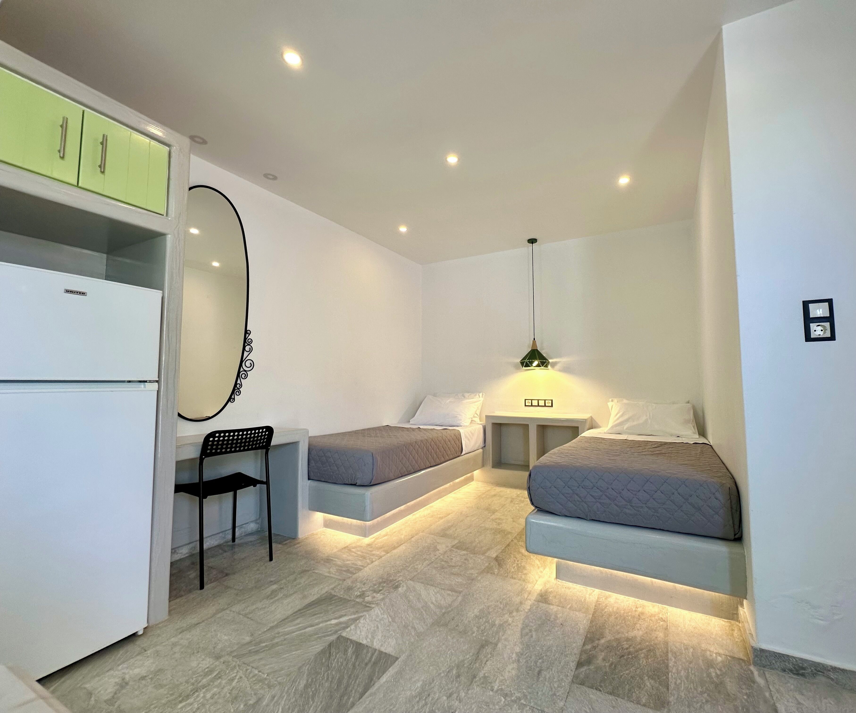 Een kleine kamer met twee bedden, een bureau, een stoel, een spiegel en een koelkast.