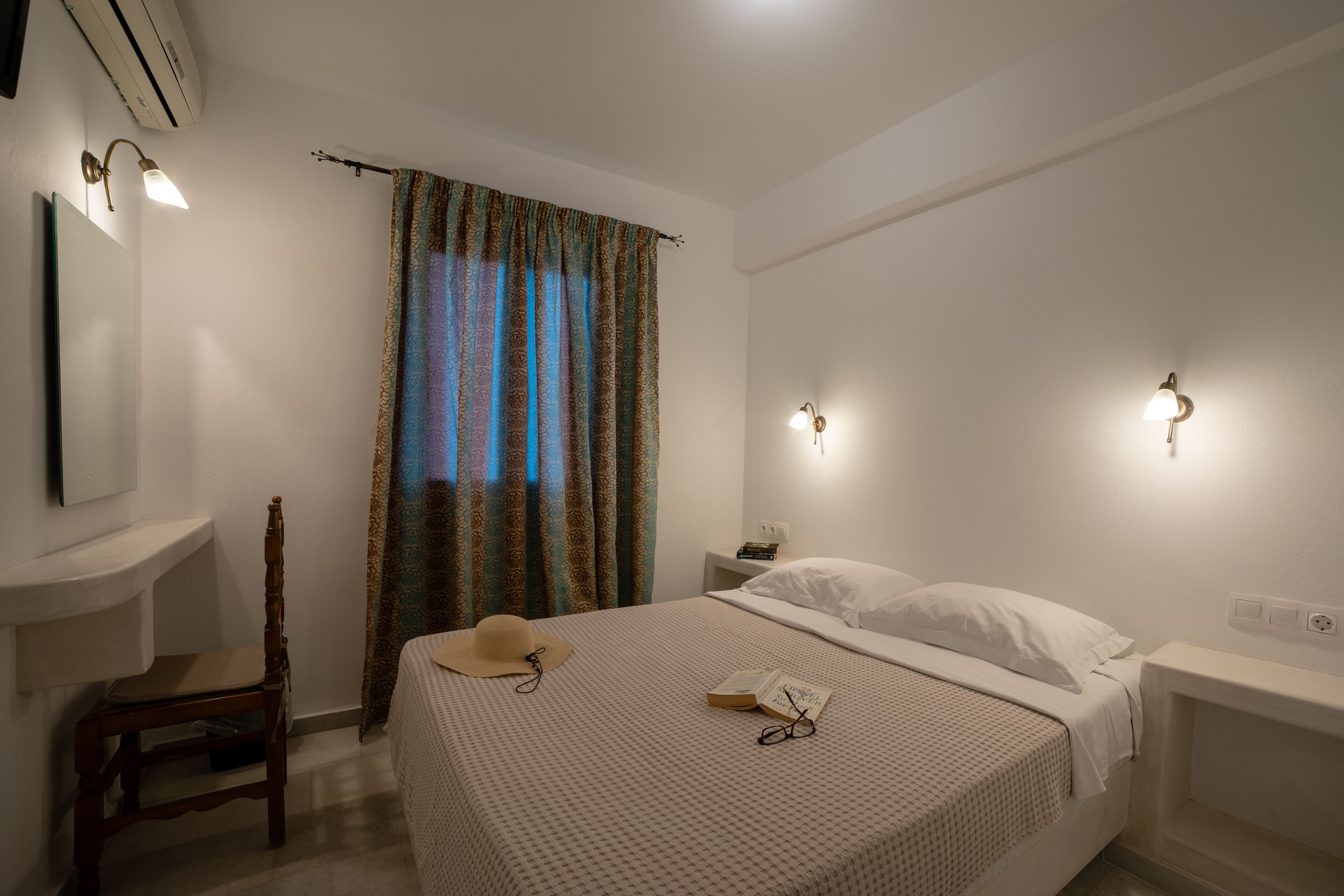 Un dormitorio con cama, una silla, una mesita de noche, una lámpara y una ventana con cortinas.