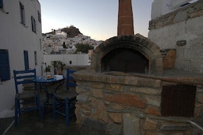 A stone-built outdoor oven with a chimney, a table with chairs, and a view of a white-washed village in the background. - Aphrodite Hotel & Apartments (Ios)