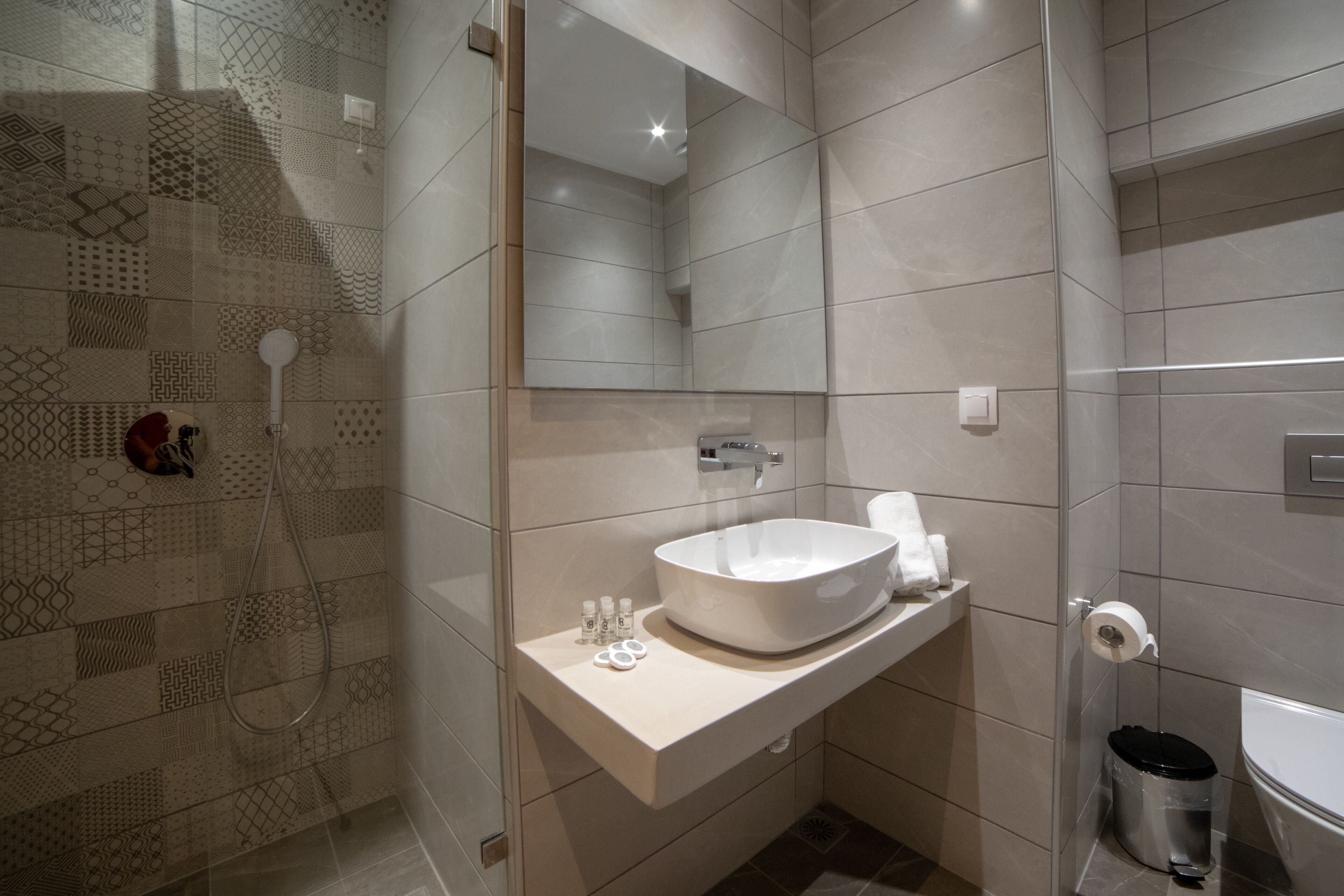Un baño moderno con un espejo grande, un lavamanos con una pila y una zona de ducha con un muro de azulejos estampados.