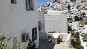 A white building with a stone-paved courtyard, a small table and chair, and a view of a hillside town with white buildings. - Aphrodite Hotel & Apartments (Ios)