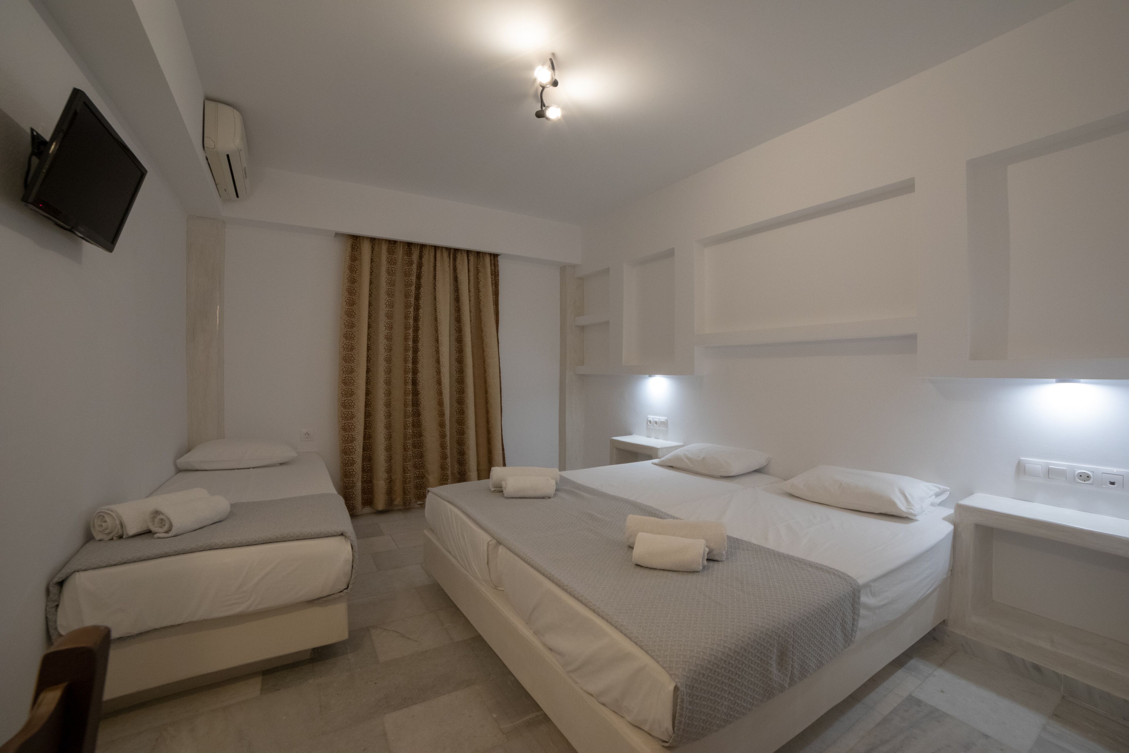 A hotel room with two beds, a TV, an air conditioning unit, and a window with curtains.