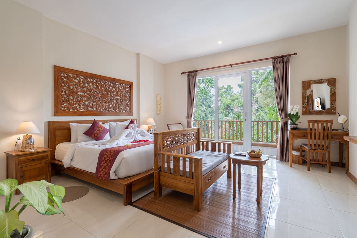 A well-lit bedroom with a bed, bedside tables, a wooden chair, a desk, and a balcony with a view of greenery.