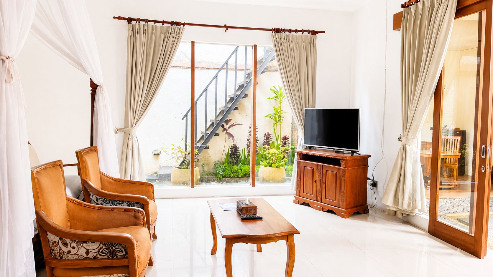 A living room with a sofa, a coffee table, and a TV on a wooden cabinet. There is a sliding glass door leading to a balcony with plants.