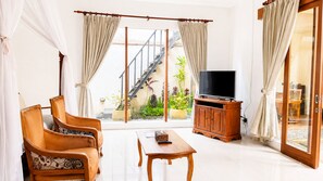 A living room with a sofa, a coffee table, and a TV on a wooden cabinet. There is a sliding glass door leading to a balcony with plants. - Ashoka Tree Resort at Tanggayuda, Ubud (Ubud)