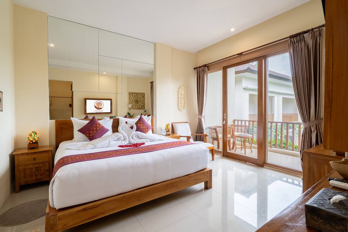 A spacious bedroom with a large bed, a wooden nightstand, a desk, and a balcony with a view of the outdoors.