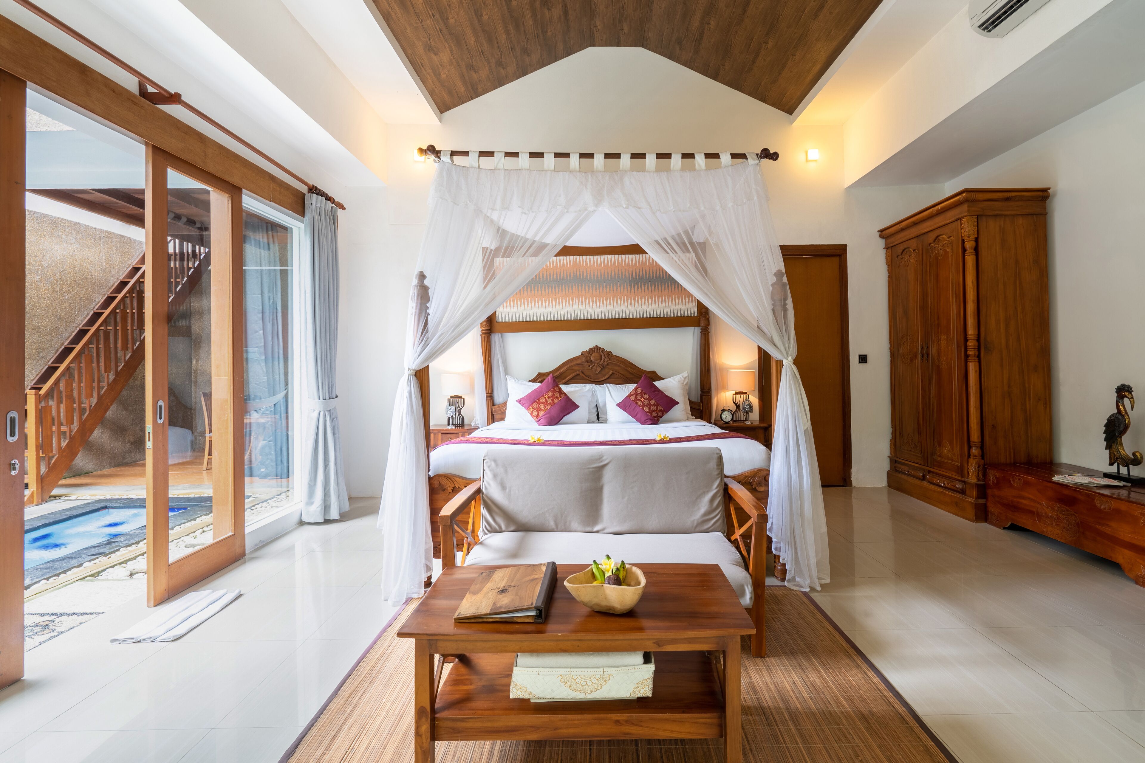 A bedroom with a large bed, a wooden table, and a view of a pool.