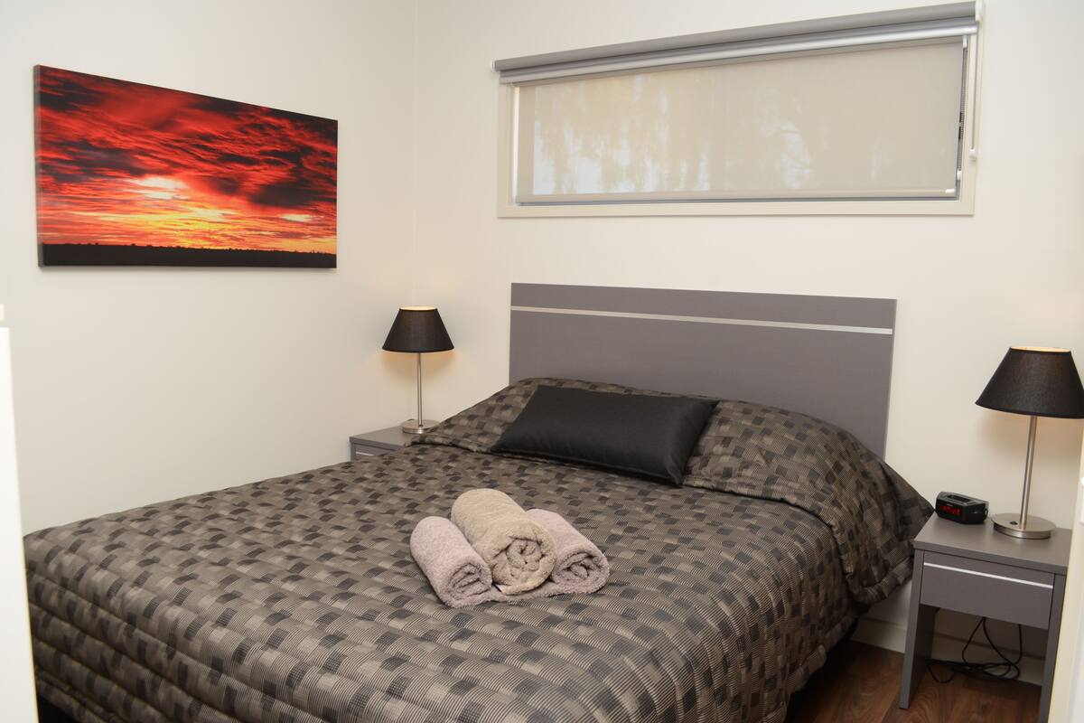 A neatly made bed with a patterned bedspread, two bedside lamps, a nightstand, and a painting of a sunset.