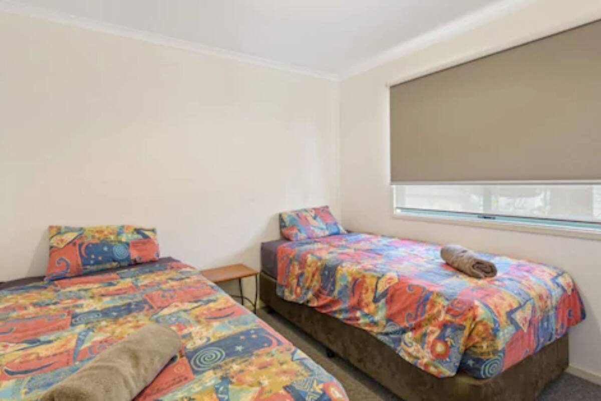 A small room with two beds, each with colorful bedding. There is a window with a blind partially drawn, and a small table between the beds.