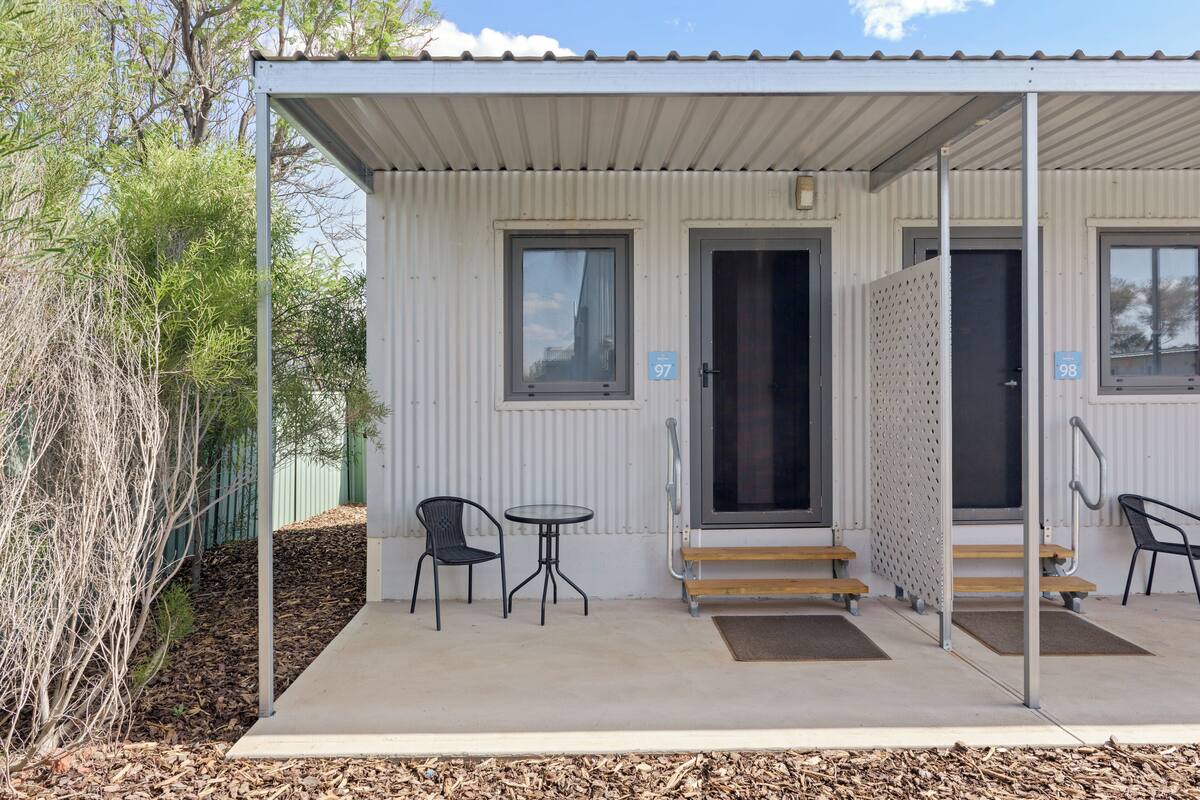 A covered outdoor area with a metal roof, two windows, a door, and a small table with a chair.