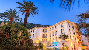 A large, classical building with multiple floors and balconies, surrounded by palm trees and greenery. - Hôtel El Paradiso (Menton)