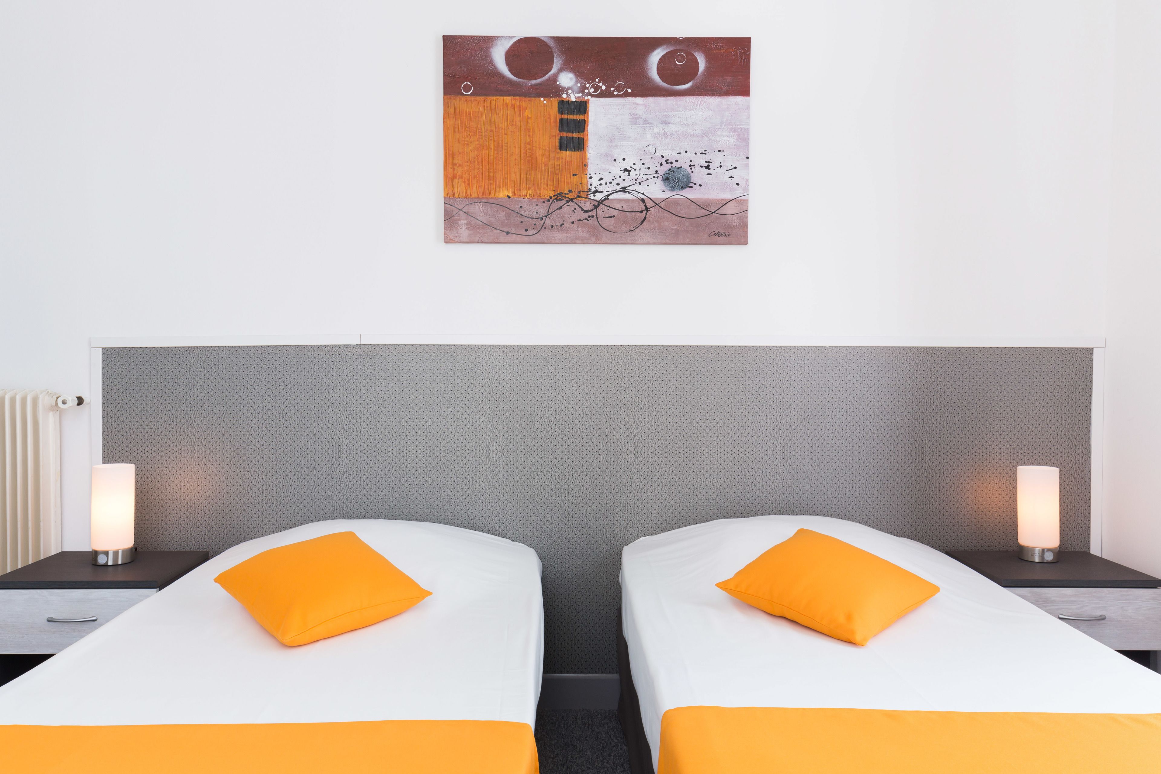 Une chambre d’hôtel avec deux lits, chacun agrémenté d’un oreiller orange, d’une tête de lit grise et d’un tableau abstrait accroché au mur.