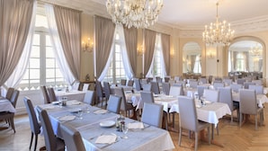 A formal dining room with multiple tables, elegant chandeliers, and large windows with curtains. - Hôtel El Paradiso (Menton)