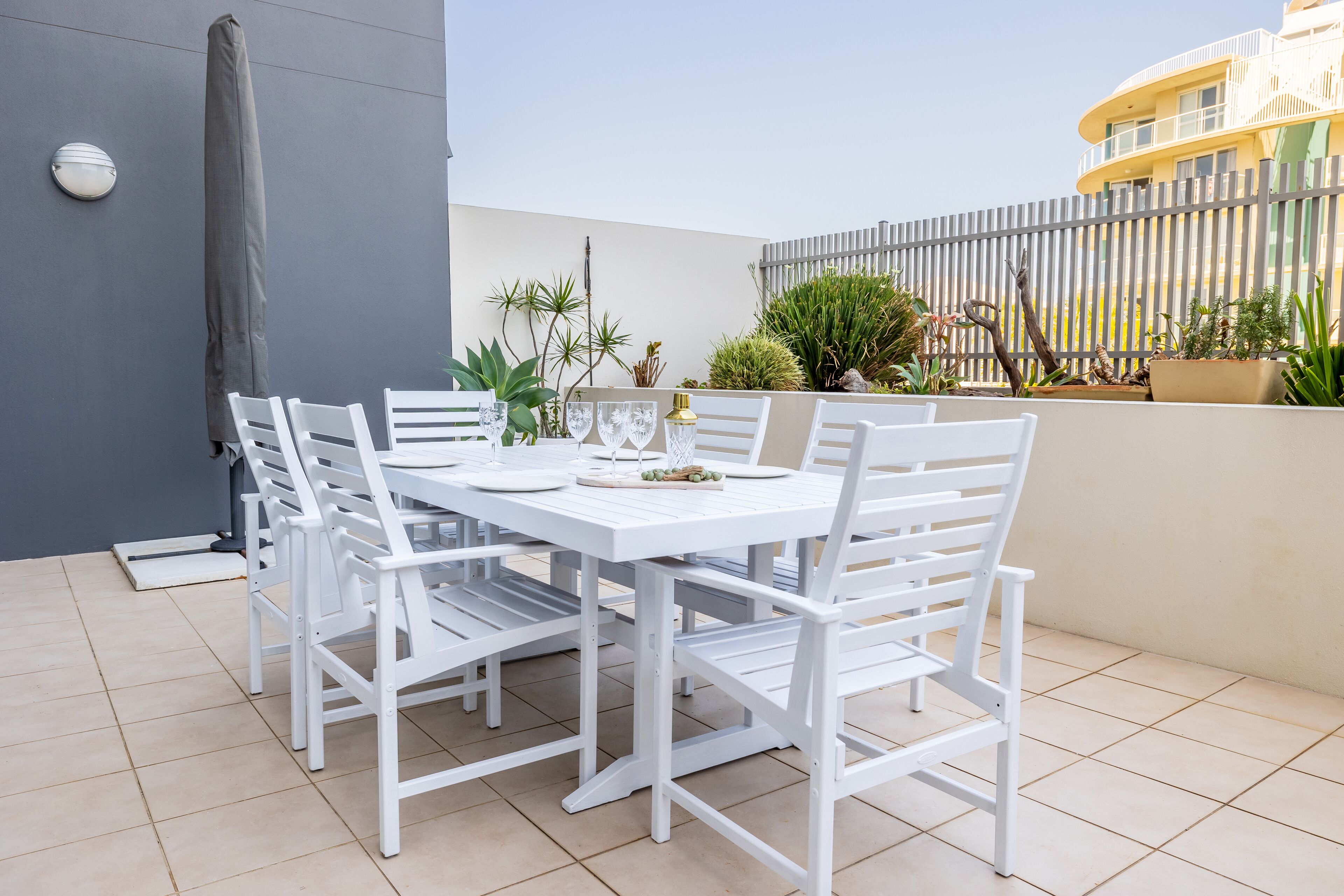 Un espace extérieur aménagé pour les repas, avec une table et des chaises, le tout dans des tons blancs, et entouré de plantes en pot et d’une clôture grise.