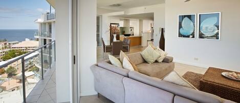 A modern living room with a sofa, a dining table, and chairs. There is a balcony with a view of the ocean.