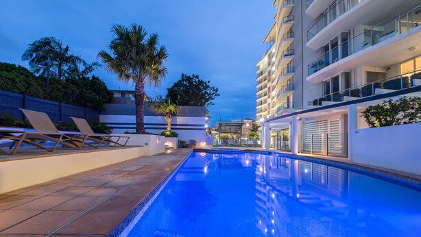 En swimmingpool med liggestole og en palme, omgivet af moderne bygninger.