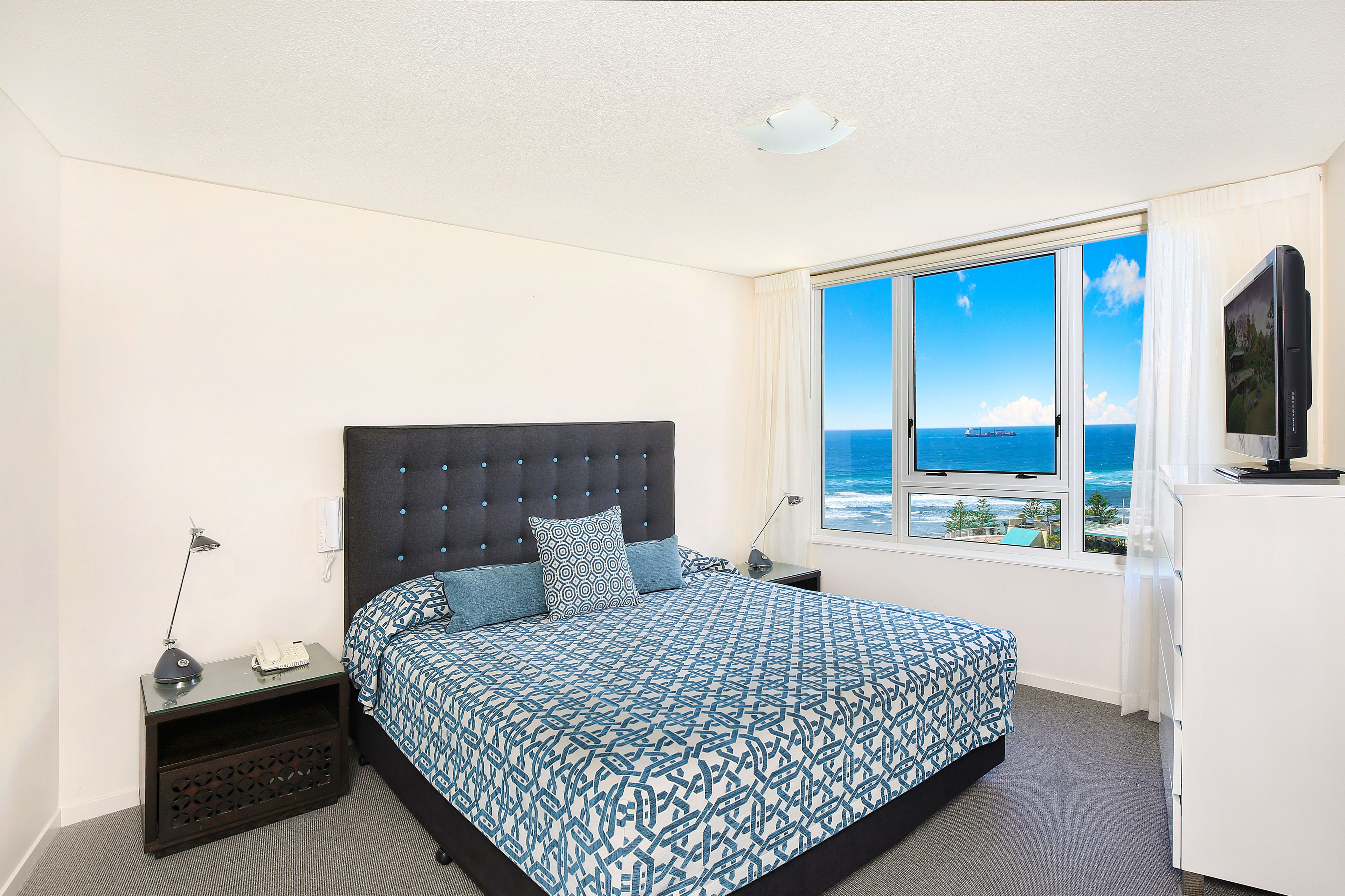 A bedroom with a large bed, a nightstand, a desk, a television, and a window with a view of the sea.