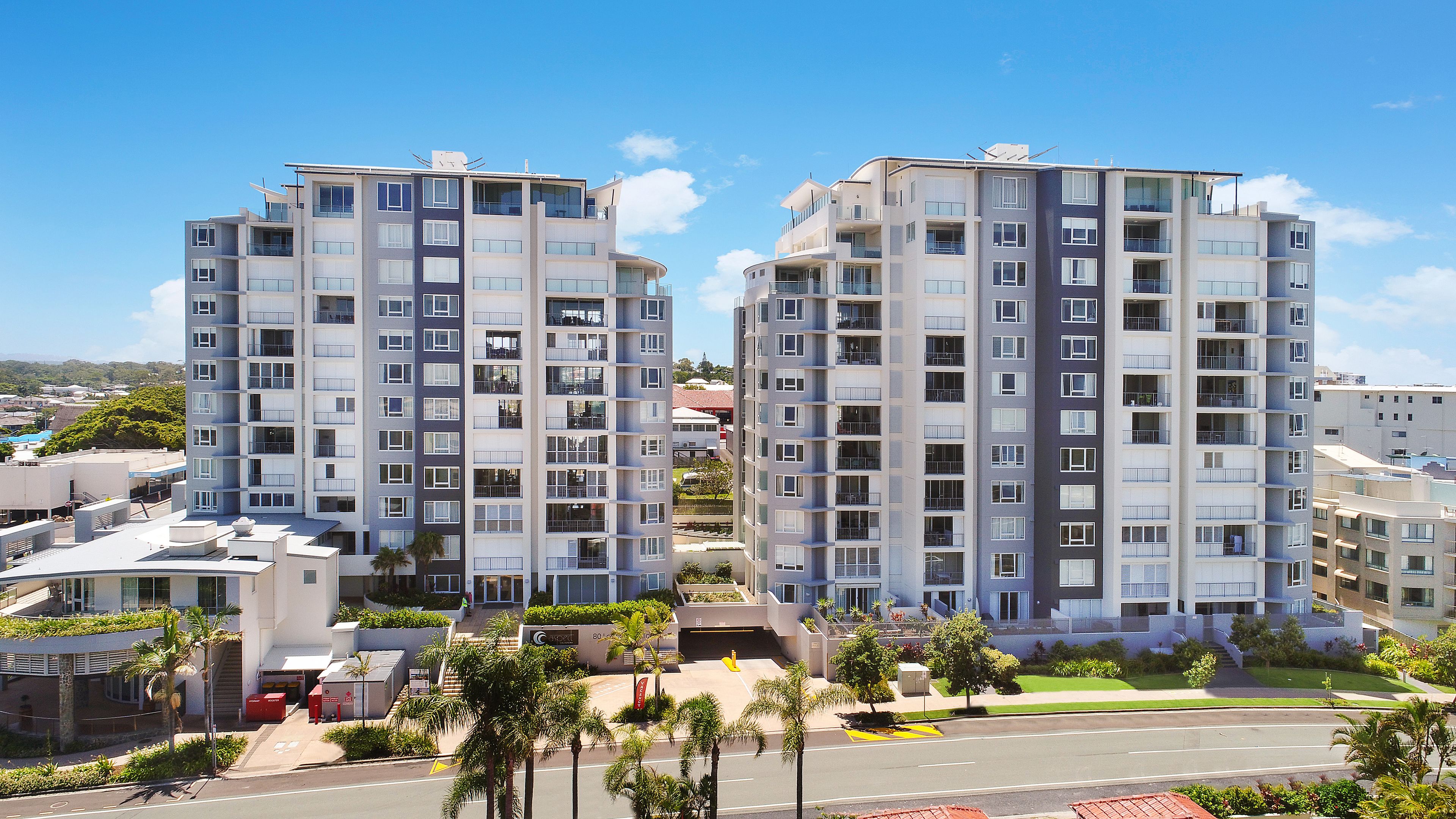 A high-rise residential building with multiple balconies, a rooftop garden, and a well-maintained surrounding area with palm trees and a clear sky.