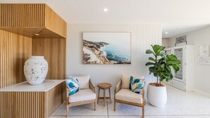 Et moderne receptionområde med træpaneler, en stor vase, to sofaer med puder, et lille rundt bord, en potteplante og et indrammet maleri af et kystlandskab.