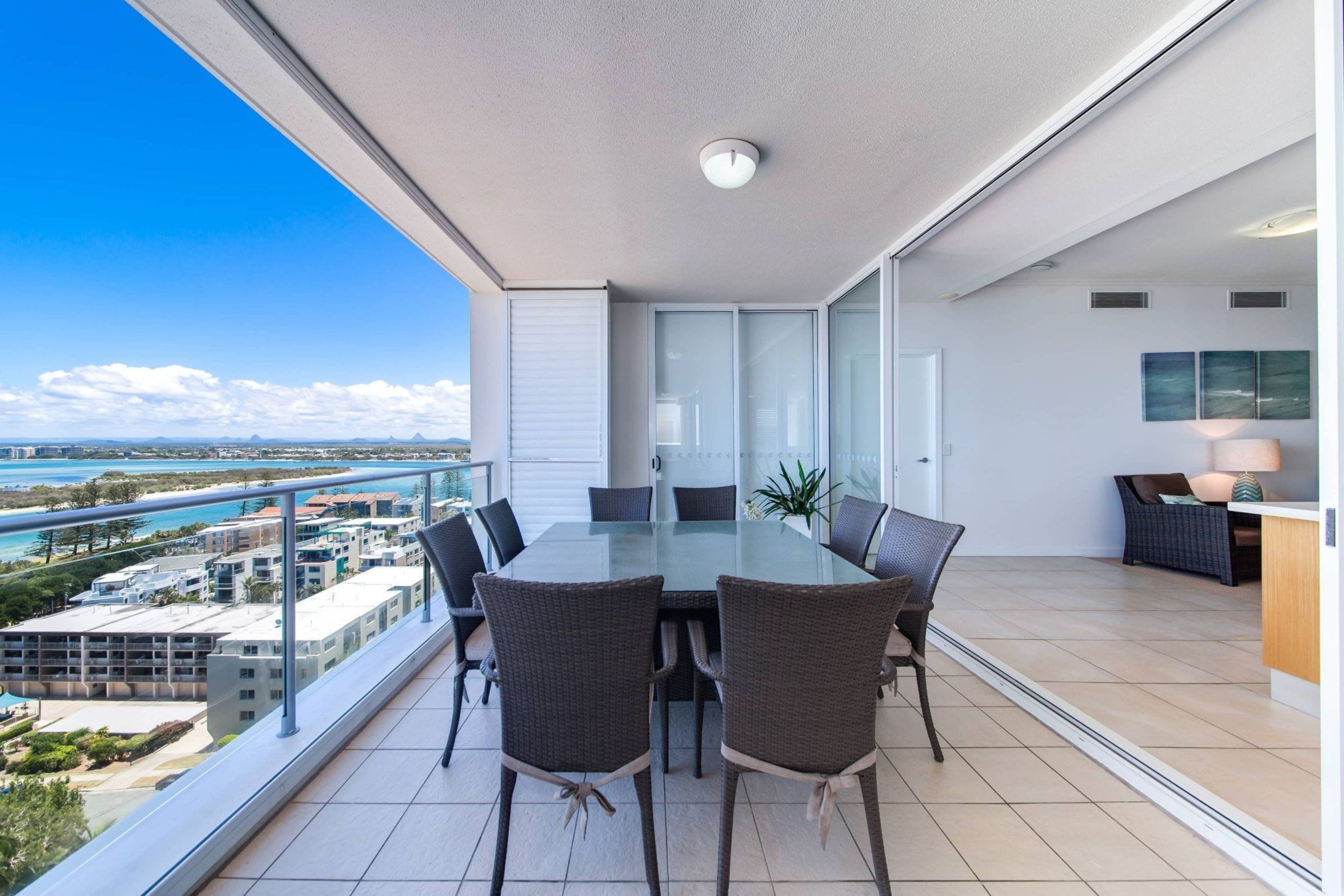 A balcony with a glass table and chairs, overlooking a coastal cityscape.