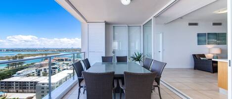 A balcony with a glass table and chairs, overlooking a coastal cityscape.
