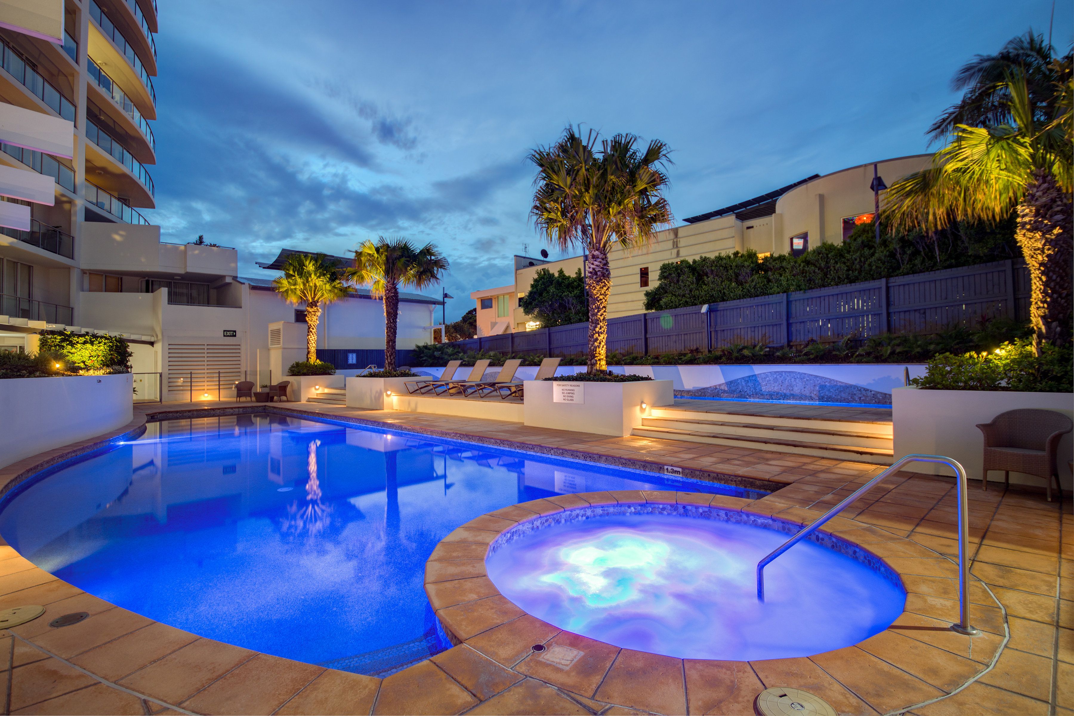 Una piscina con hidromasaje y sillones de descanso, rodeada de palmeras y un edificio con balcones.