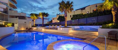 Una piscina con hidromasaje y sillones de descanso, rodeada de palmeras y un edificio con balcones.