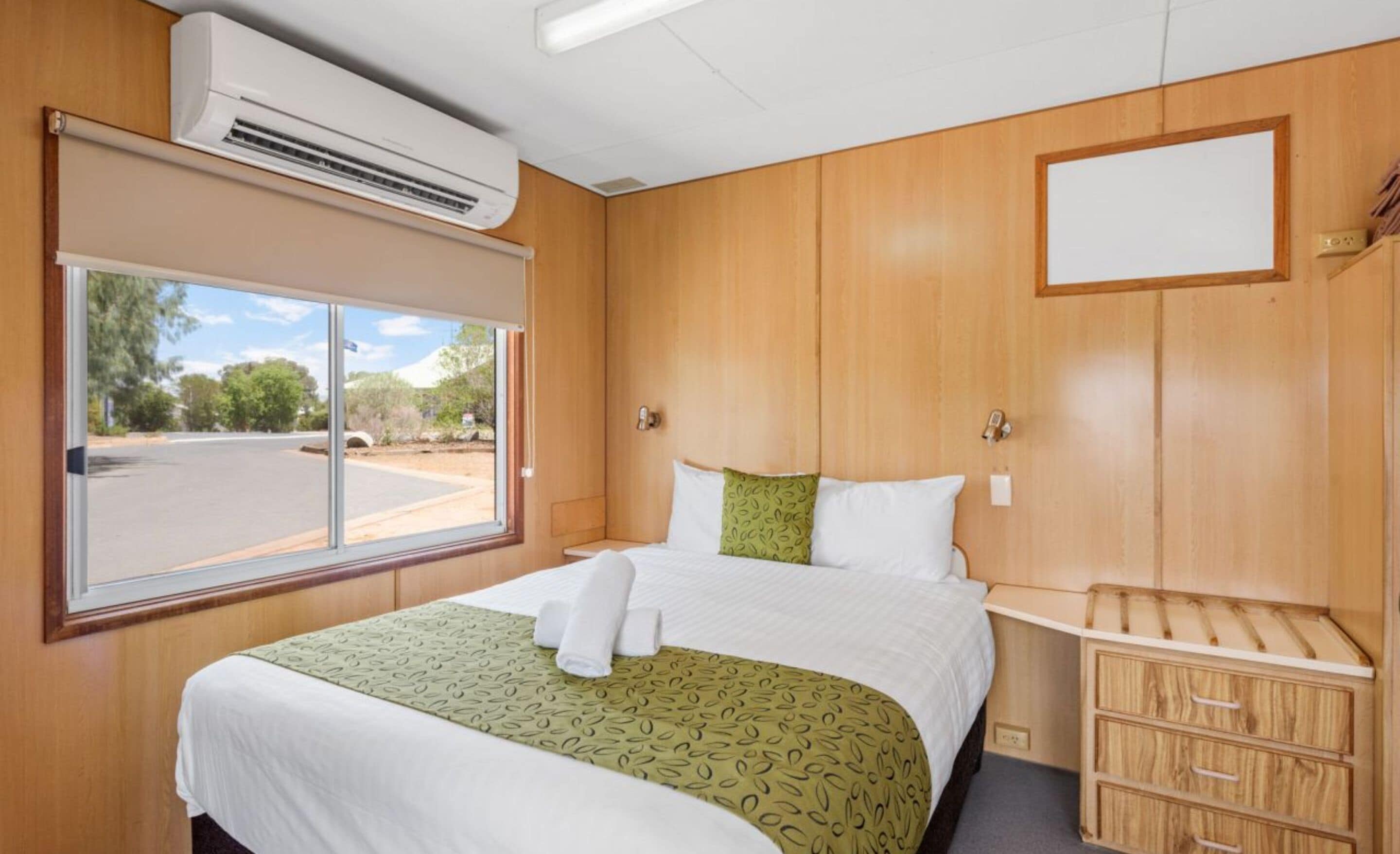 Una habitación de hotel con un ventanal grande, una cama con cubrecama verde y blanco, paneles de madera y un pequeño armario de madera.