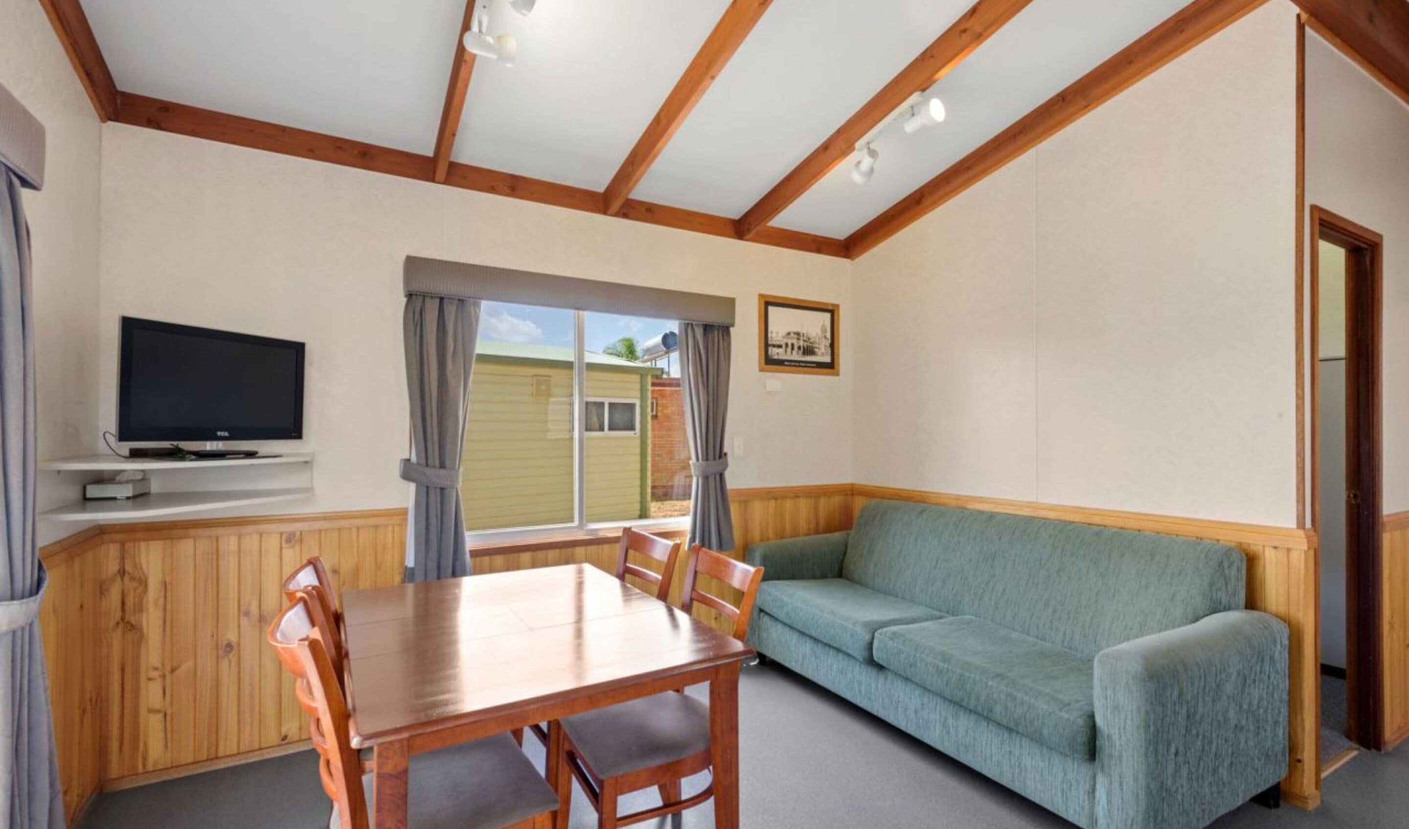Et værelse med et træspisebord, stole, en sofa, et fjernsyn på en hylde og et vindue med gardiner.