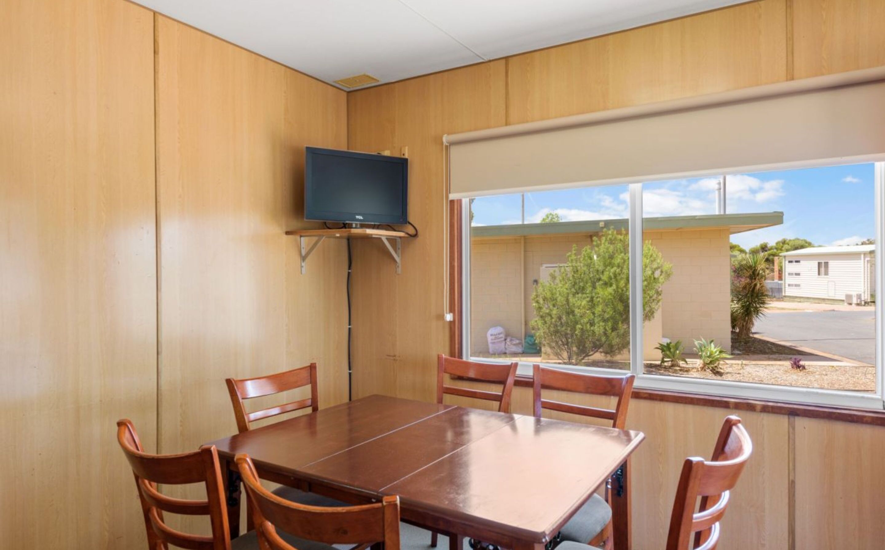 A room with a wooden interior, a TV mounted on the wall, a dining table with chairs, and a view of the outside through a large window.