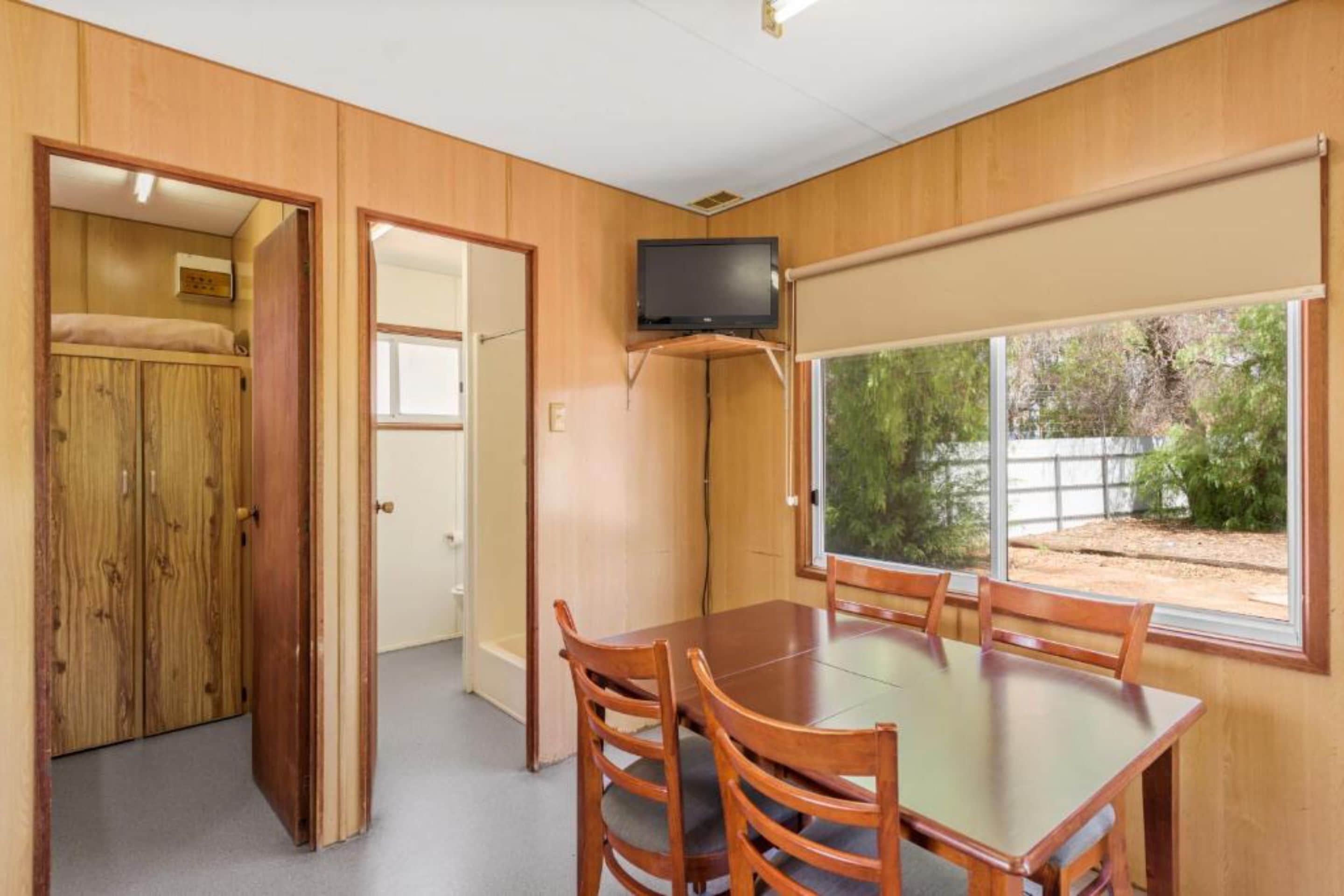 Een kamer met een houten vloer, een eettafel met stoelen, een aan de muur bevestigde televisie en een raam met uitzicht op groen.