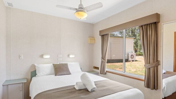 Habitación de hotel con cama, ventilador de techo, ventana con cortinas y unidad de aire acondicionado en el exterior.