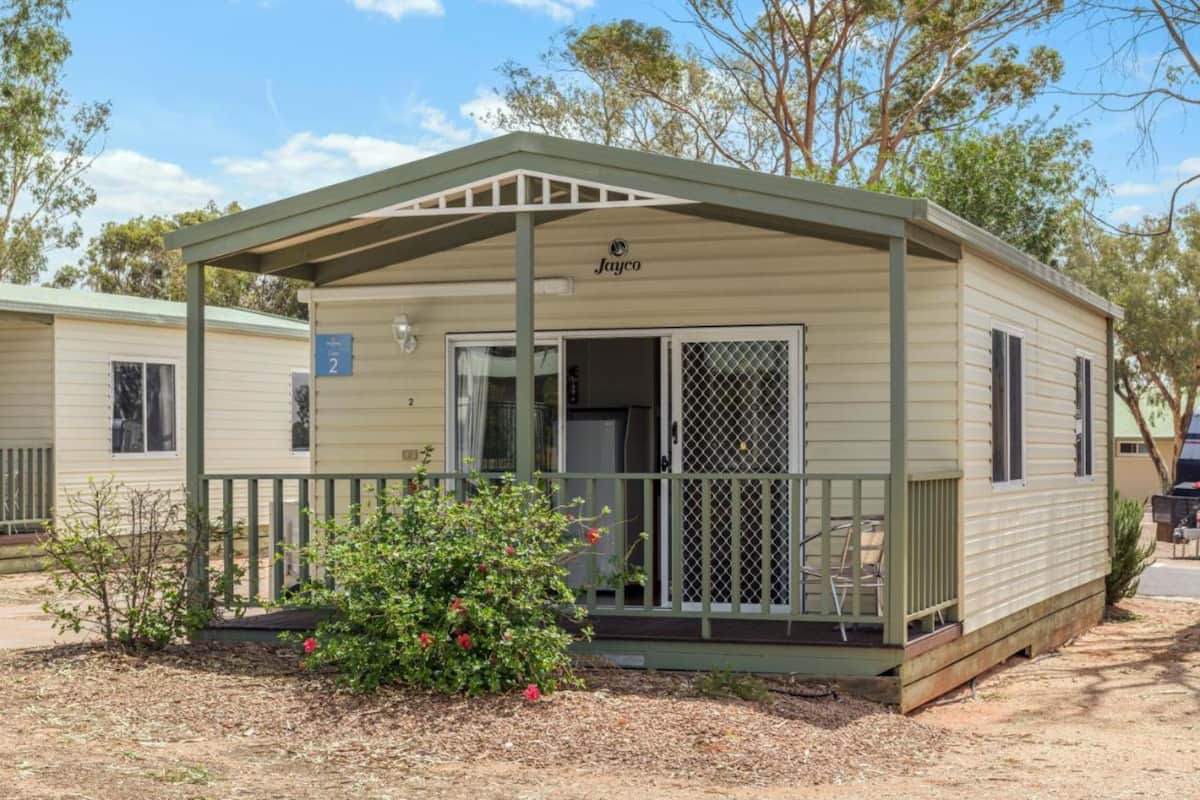 A mobile home with a covered porch, a small garden with red flowers, and a sign indicating the unit number.