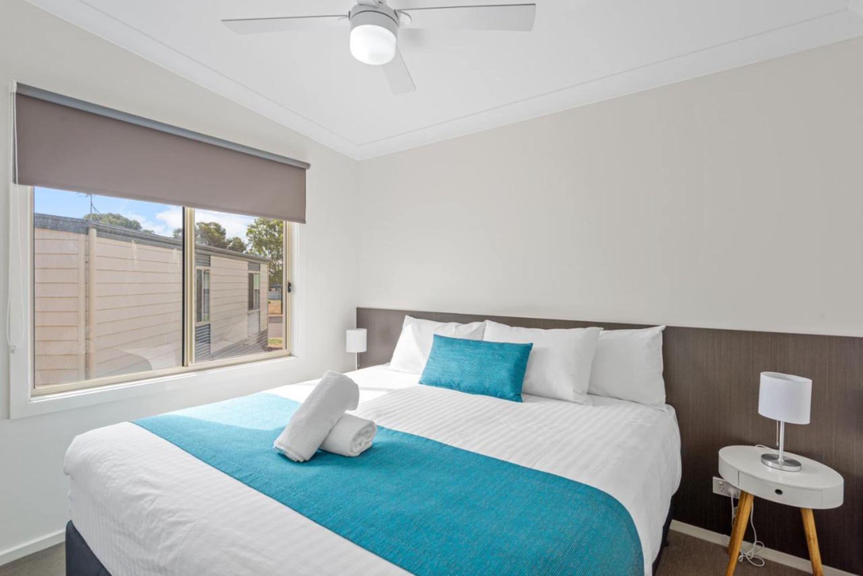 A hotel room with a large bed, a ceiling fan, a window with blinds, and a bedside table with a lamp.
