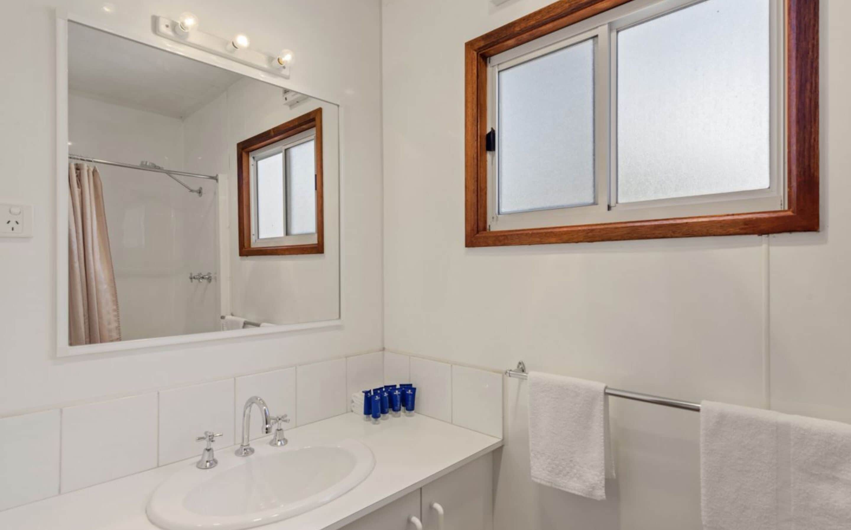 A bathroom with a large mirror, a sink with two faucets, and a window above the mirror.