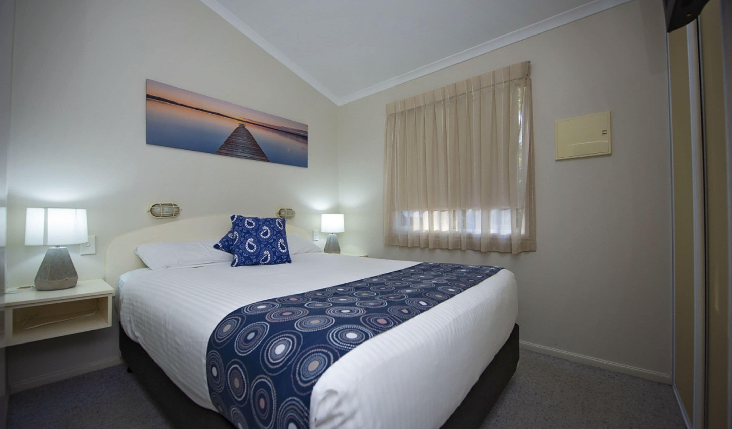 A hotel room with a bed, two bedside tables with lamps, a painting on the wall, and a window with curtains.