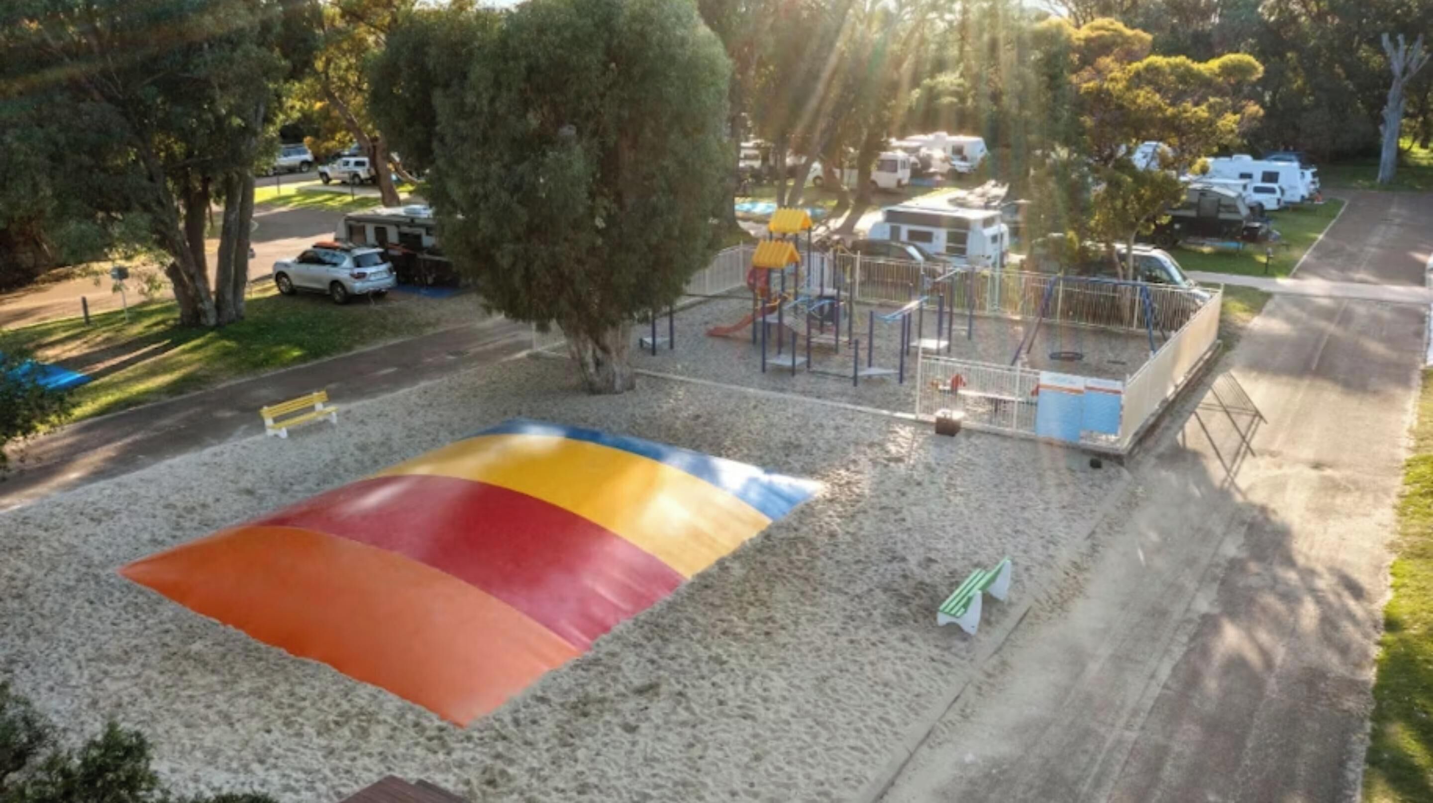 A playground with a colorful slide and a fenced area, surrounded by trees and parked vehicles.